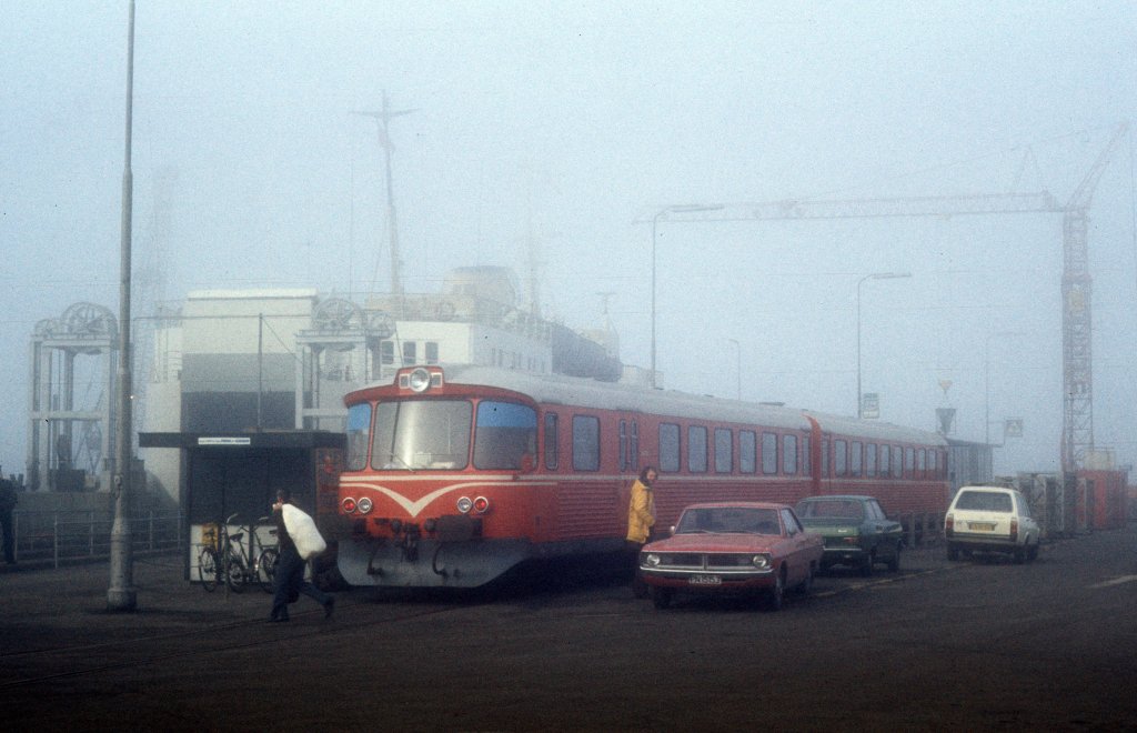 HP, Hjørring Privatbaner, deren Züge die Bahnstecke Hjørring - Hirtshals in Nordjütland bedienten: Im Fährhafen von Hirtshals hält am Morgen des 24. Februar 1975 ein  Y-Zug  bestehend aus einem Triebwagen (Ym) und einem Steuerwagen (Ys). - Einige Züge der HP fuhren zwischen dem HP-Bahnhof in Hirtshals und dem Fährhafen, um Fahrgäste zur / von der Fähre zwischen Hirtshals und Kristianssand in Norwegen zu bringen. Wegen des Morgennebels sieht man nur undeutlich das Fährschiff, das im Fährbett liegt. 