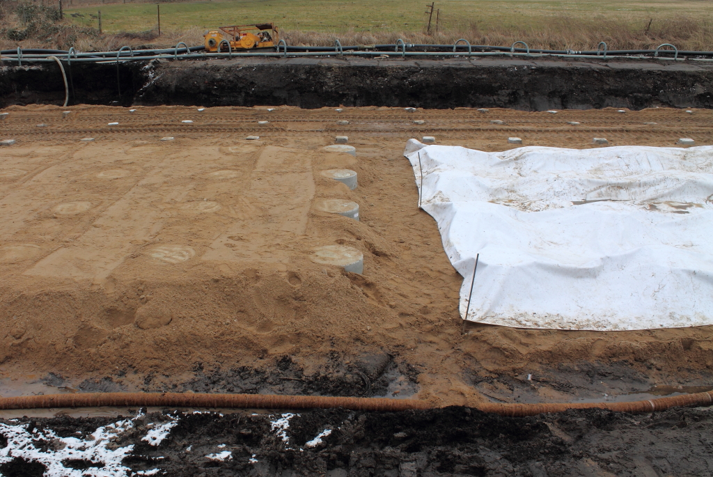 Neues von der Baustelle in Nassenheide am 08.02.2013. Hier wird gegen Moor, Grundwasser und Klte gekmpft. Ganz vorn Drnage und im Hintergrund Grundwasserabsenkung, die schon seit September pausenlos luft. Links im Bild haben die Rammpfhle ein  Htchen  zwecks Druckverteilung erhalten. Rechts in der Bildmitte: Um den Kraftschluss zwischen Htchen und Rammpahl herzustellen, wird dnnflssiger Beton eingefllt und mit Warmluft beheizt. Dahinter die noch unbehandelten Rammpfhle. ber der rechten Seite wird der Damm in Geo-Gitter-Bauweise errichtet. Also Fortsetzung  Neues von der Baustelle  folgt....