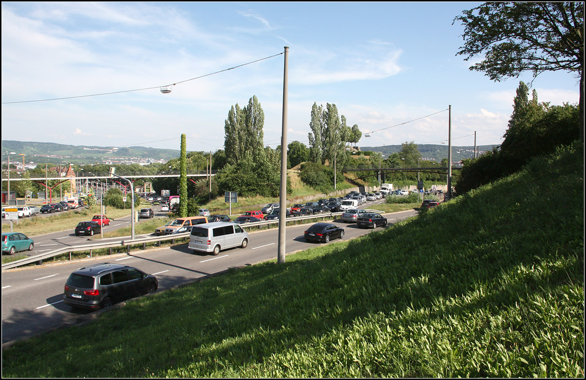 . Ziemlich umgekrempelt und die Bahnen im Tunnel -

Blick in die Pragstraße (links) und die Heilbronner Straße (rechts) vom Stuttgarter Pragsattel aus. Im Vergleich zum Bild von Kurt Rasmussen hat sich sehr viel verändert:

http://www.bahnbilder.de/bild/deutschland~strassenbahn~stuttgart-keine-stadtbahn/670836/stuttgart-ssb-sl-5-gt4-699.html

Die Bebauung zwischen den Bundesstraßen ist verschwunden, auch deren Lage hat sich nach Nordosten verschoben. Wahrscheinlich stand das Gebäude, dort wo ungefähr in Bildmitte heute die Autos fahren. Dadurch ist die Heilbronner Straße von der Wohnbebauung, die sich rechts außerhalb des Bildes befindet und auf Kurts Bild erkennbar ist, abgerückt. Auch wenn es nicht so aussieht sind unsere Fotostandpunkte möglicherweise ziemlich nah beieinander, meiner etwas höher. Die SSB-Bahnen fahren seit 1990 im Tunnel. Die Rampen liegen jeweils im Bereich der beiden Fußgängerbrücken. Das Gelände rechts ist jetzt recht hügelig, auch um dem Wohngebiet auf dieser Seite Lärmschutz zu bieten. Auf der gegenüberliegenden Seite hier in Bildmitte entstand zur IGA '93 ein Park mit Aussichtshügel. Die Bundesstraße 10 (Pragstraße) befindet sich seit 2006 im Tunnel.

27.06.2016 (M)