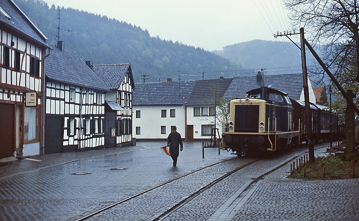 212 070-7 überquert an einem Frühjahrsmorgen 1988 den Dorfplatz in Olef (Strecke Kall - Hellenthal). Der Rangierer ging dem Zug voraus und sicherte mit der Fahne den Fahrweg. 