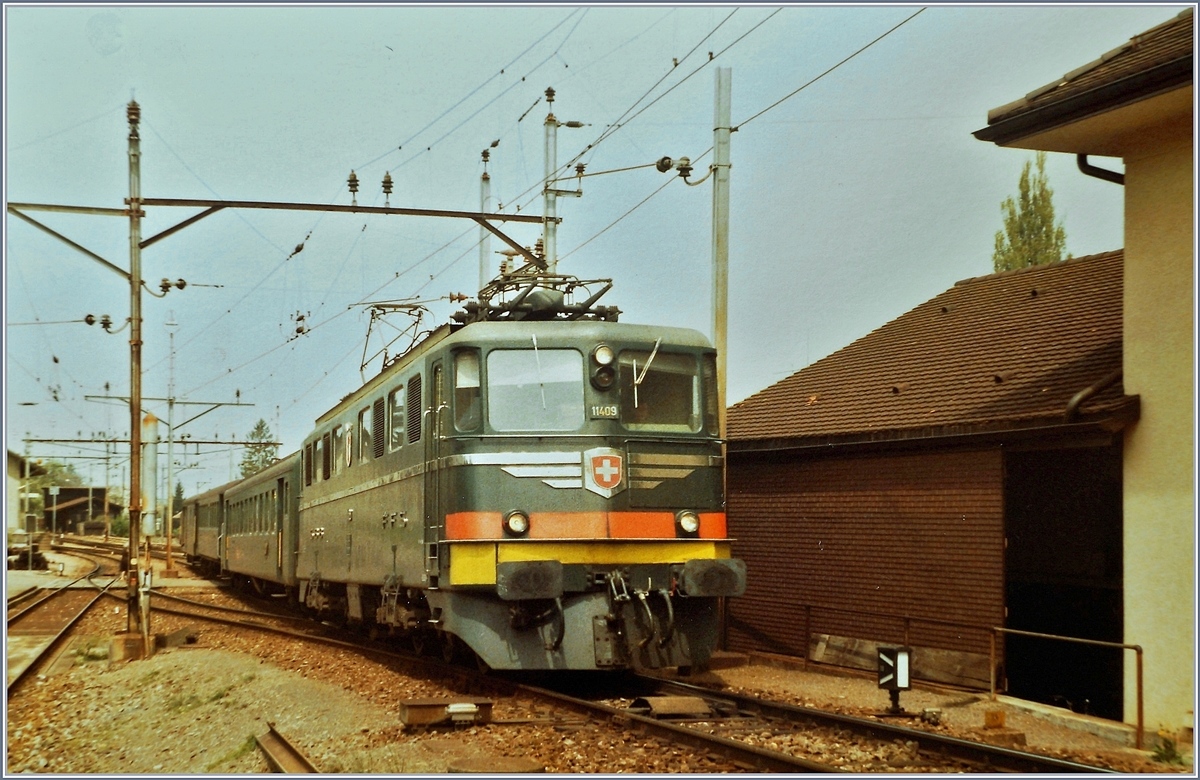 Anstelle eines ausgefallenen De 4/4 nimmt die Ae 6/6 11409 mit einem Regionalzug nach Beromünster die recht steile Rampe hinter dem Bahnhof in Angriff. In Hintergrund sichtbar: die Gleisanlage der Spitzkehre in Beinwil am See. 
16. Mai 1984.