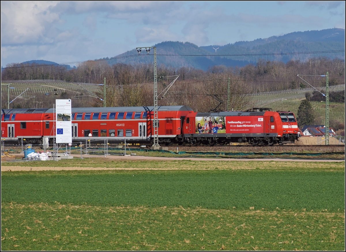 Bahn im Markgräflerland. Mit Baden-Württemberg-Ticket-Werbung an der Baustellenankündigung für den viergleisigen Ausbau vorüber. 146 113-6 als echte Rheintallok ist das Zugpferd an diesem Regionalexpress nach Basel. Müllheim, März 2019.