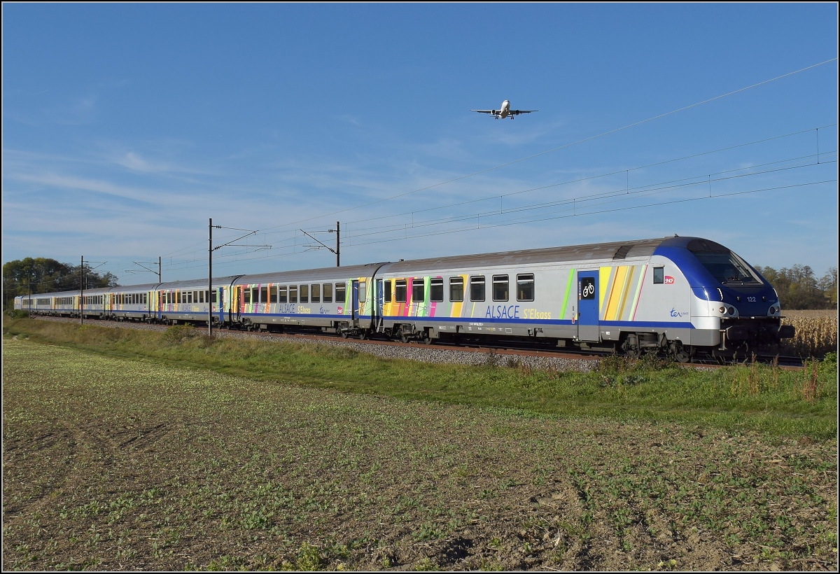 Bahn im südlichen Elsass. TER 96263 mit Schiebelok Sybic 26143 gewinnt beim Landeanflug auf Basel gegen den Airbus A320-200 TC-DCJ der Pegasus Air. Bartenheim, Oktober 2017.