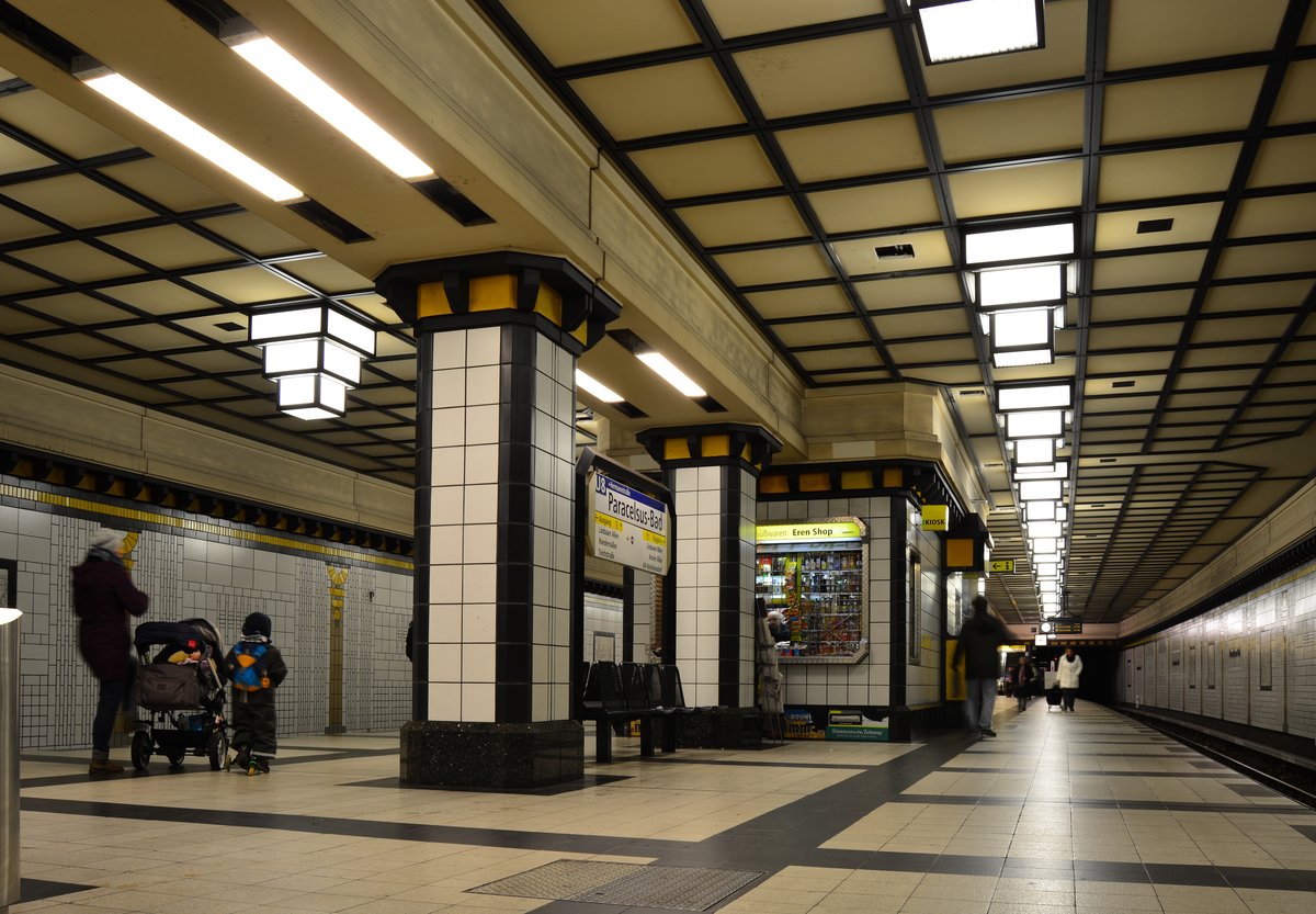 Blick durch den am 27.April 1987 eröffneten U-Bahnhof Paracelusbad. Entworfen wurde der Bahnhof von Rainer G. Rümmler. Die Wände erinnern in stilisierter Form durch die weiße sanitärfliesenartige Wandgestaltung an Schwimmbäder, genauso wie die Säulen, die auf den Wänden abgebildet sind, an römische Badeanlagen erinnern. Die Decke ist mit einem quadratischen Muster aus Holzbalken bedeckt, die Säulen in der Mitte des Bahnsteigganges ähneln denen in einem Schwimmbad. Ein Augenmerk sind die Lampen an der Decke, die Tropfsteine verkörpern und aus Quadern im Stil des Art Déco bestehen.

Berlin Reinikendorf 03.01.2018


Seite „U-Bahnhof Paracelsus-Bad“. In: Wikipedia, Die freie Enzyklopädie. Bearbeitungsstand: 22. Juni 2017, 17:22 UTC. URL: https://de.wikipedia.org/w/index.php?title=U-Bahnhof_Paracelsus-Bad&oldid=166627705