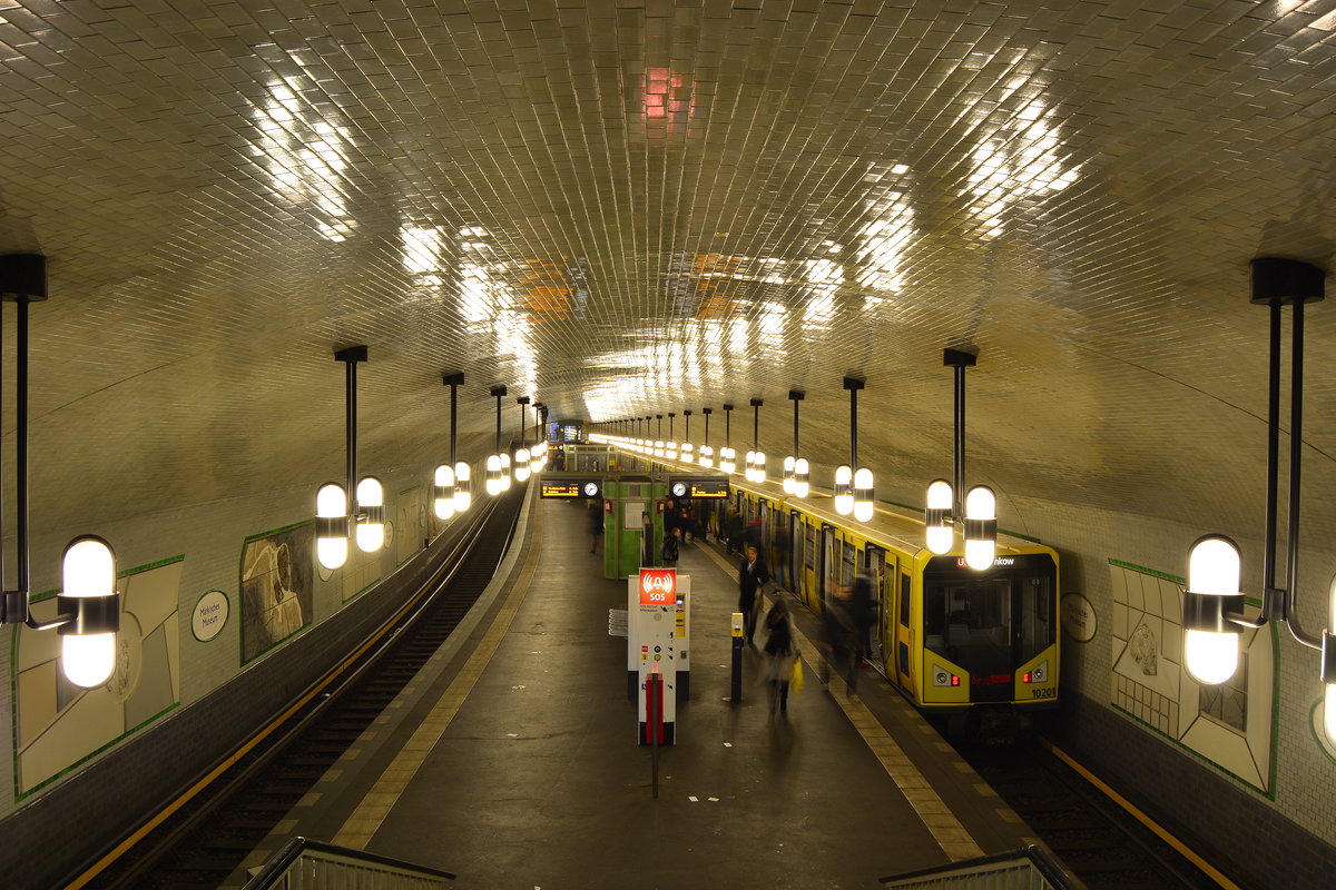 Blick über die am 1.Juli 1913 eröffnete U-Bahn Station Märkisches Museum. Rechts steht Triebzug 1020 der Baureihe H als U2 nach Pankow.

Planung und erste Jahrzehnte

Im Zuge der Verlängerung der damals sogenannten Spittelmarktlinie als Centrumslinie zum Alexanderplatz entstand der Bahnhof Inselbrücke in unmittelbarer Ufernähe der im anschließenden Streckenverlauf zu querenden Spree, weswegen er mit für damalige Verhältnisse sehr tiefen sechseinhalb Metern unterhalb des Straßenniveaus eine bauliche Besonderheit jener Zeit darstellte.

Dank der tiefen Lage konnte Alfred Grenander als Architekt die Station mit einer großen und geräumigen Halle und einem Korbbogengewölbe ausstatten. Aufgrund dieser in Berlin ungewöhnlichen Bauweise wurde die Station oft mit jenen der Pariser Métro in Verbindung gebracht. Im Berliner U-Bahn-Netz verfügt nur der U-Bahnhof Platz der Luftbrücke über eine solche stützenlose Konstruktion.

Der Mittelbahnsteig ist 121,3 Meter lang und 7,6 Meter breit; die Halle ist an ihrer größten Stelle 5,2 Meter hoch.

Für den Bahnhof wählte Grenander die Kennfarbe Grün, ebenso wie für den Bahnhof Leipziger Platz, denn sein Farbschema für diese Strecke sah eine regelmäßige Wiederholung der Farben vor. Der Bahnhof, der jeweils in östlicher und westlicher Richtung ein kleines Mittelgeschoss besitzt, erhielt einen 121,3 Meter langen und 7,6 Meter breiten Mittelbahnsteig.
Der U-Bahnhof Inselbrücke im Jahre 1913 (seit 1935: Märkisches Museum)

Nach langwierigen Bauarbeiten nahm die Hochbahngesellschaft am 1. Juli 1913 den Betrieb auf der 1,7 Kilometer langen Linie Spittelmarkt – Alexanderplatz auf.

Seit 1928 trugen die U-Bahn-Linien, die den Bahnhof Inselbrücke befuhren, die Bezeichnungen AI und AII.

Seinen heutigen Namen erhielt der Bahnhof 1935 auf Antrag der Leitung des in der Nähe liegenden Märkischen Museums.


Zweiter Weltkrieg

Am 24. Mai 1944 erlitt die Gewölbedecke durch eine Fliegerbombe leichte Schäden. Anfang April 1945 drang Wasser in Folge eines Bombentreffers in den nahegelegenen Klostertunnel sowie auf Umwegen vom in den letzten Kriegstagen gesprengten S-Bahn-Tunnel unter dem Landwehrkanal ein und drang bis zum Bahnhof vor. Zu diesem Zeitpunkt war der Betrieb allerdings längst eingestellt, da kein Strom mehr zur Verfügung stand.


Nachkriegszeit

Am 31. Juli 1945 wurde der Bahnhof Märkisches Museum wieder in Betrieb genommen. Er diente zunächst als Endstation eines Pendelverkehrs zum Bahnhof Stadtmitte. Bis zum 1. November 1945 konnte auch die Spreeunterführung zum U-Bahnhof Klosterstraße vorerst eingleisig wiederhergestellt werden. Am 15. September 1946 war der Umlaufbetrieb zwischen Pankow und Ruhleben zumindest bis zur durch den Mauerbau bedingten Unterbrechung der Linie wieder vollständig.


DDR-Zeit


Die DDR stellte die Instandhaltung des Bahnhofs lange Zeit zugunsten eines Ausbaus der Verkehrsverbindungen in die entstehenden Berliner Neubaugebiete zurück. Erst zur 750-Jahr-Feier Berlins wurde ein Sanierungsprogramm beschlossen. In diesem Zusammenhang nahmen die Künstler Jo Doese (Materialcollagen) sowie Karl-Heinz Schäfer und Ulrich Jörke (24 Stuckreliefs) gestalterisch Bezug auf das benachbarte und namensgebende Museum. Die Autorin Ingrid Bartmann-Kompa unterstrich dies mit passenden Zitaten.[9] Außerdem wurde eine neue Lichtanlage, bestehend aus kugelförmigen Pendelleuchten, installiert.

Bei dieser Umgestaltung konnte (und musste) die eigentliche Deckenstruktur ermittelt werden, da kein tragfähiger Beton für die Anbringung der Lampen vorhanden war. Das Korbbogengewölbe besteht nach diesen Erkenntnissen aus einer rechteckigen, mit mehreren Kantenbrüchen vollendeten Decke, dabei sind die Lücken mit Putz aufgefüllt. An den beiden Ausgängen ließ die Stadt Berlin neue, den historischen Vorbildern nachempfundene, Eingangsportale errichten.


Nachwendezeit

Alle diese Arbeiten änderten nichts an der Notwendigkeit einer Grundsanierung, die auch die Gewölbedecke selbst mit einschloss. Diese begann im Jahre, wobei anfangs – ohne Zustimmung der Denkmalschutzbehörde – alle Fliesen abgeschlagen wurden. Nachdem sich die Verkehrsbetriebe mit der Senatskulturverwaltung nachträglich auf eine denkmalgerechte Sanierung geeinigt hatten, investierte die BVG gemeinsam mit Land und Bund zwölf Millionen Mark in die Bauarbeiten.

Dabei wurden unter anderem

-alle Fliesen originalgetreu nachgebrannt,
-eine neue Beleuchtungsanlage installiert,
-die Bahnsteigplatte erneuert und
-die Wandcollagen aufgefrischt.

Zusätzlich baute die BVG einen Aufzug von der Mitte des Bahnsteiges zur oberirdischen Verkehrsinsel auf der Wallstraße ein. Die Arbeiten konnten im Jahr 2002 abgeschlossen werden.

Die Sanierung dieses Bahnhofs bildete einen Anfangspunkt für das von der BVG geplante „Geschichtslinienkonzept“, nach dem alle Bahnhöfe zwischen Alexanderplatz und Stadtmitte in ihren ursprünglichen Zustand zurückversetzt werden.

Berlin Mitte 03.01.2018


Seite „U-Bahnhof Märkisches Museum“. In: Wikipedia, Die freie Enzyklopädie. Bearbeitungsstand: 17. Juni 2017, 10:27 UTC. URL: https://de.wikipedia.org/w/index.php?title=U-Bahnhof_M%C3%A4rkisches_Museum&oldid=166472632     