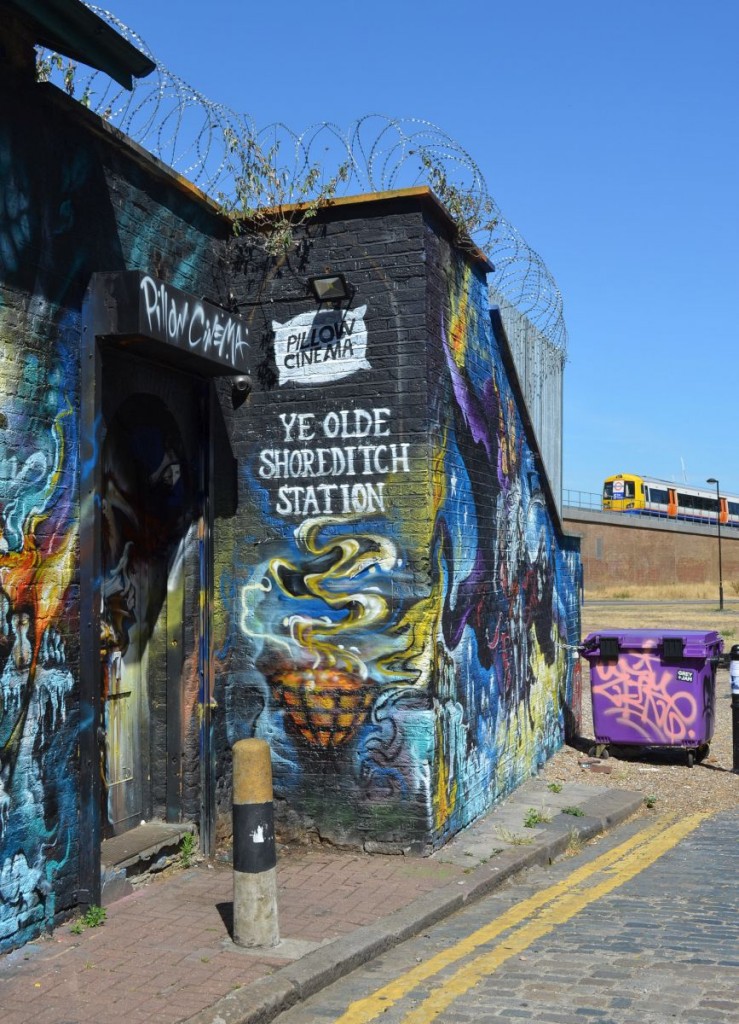 Das einstige Bahnhofsgebäude von  Shoreditch  - heute zum Kunstobjekt geworden. Es gehörte zur Underground East London Line, die jetzt von London Overground betrieben wird. Im Hintergrund nähert sich ein Zug dem neuen Bahnhof  Shoreditch High Street . 10.7.2015