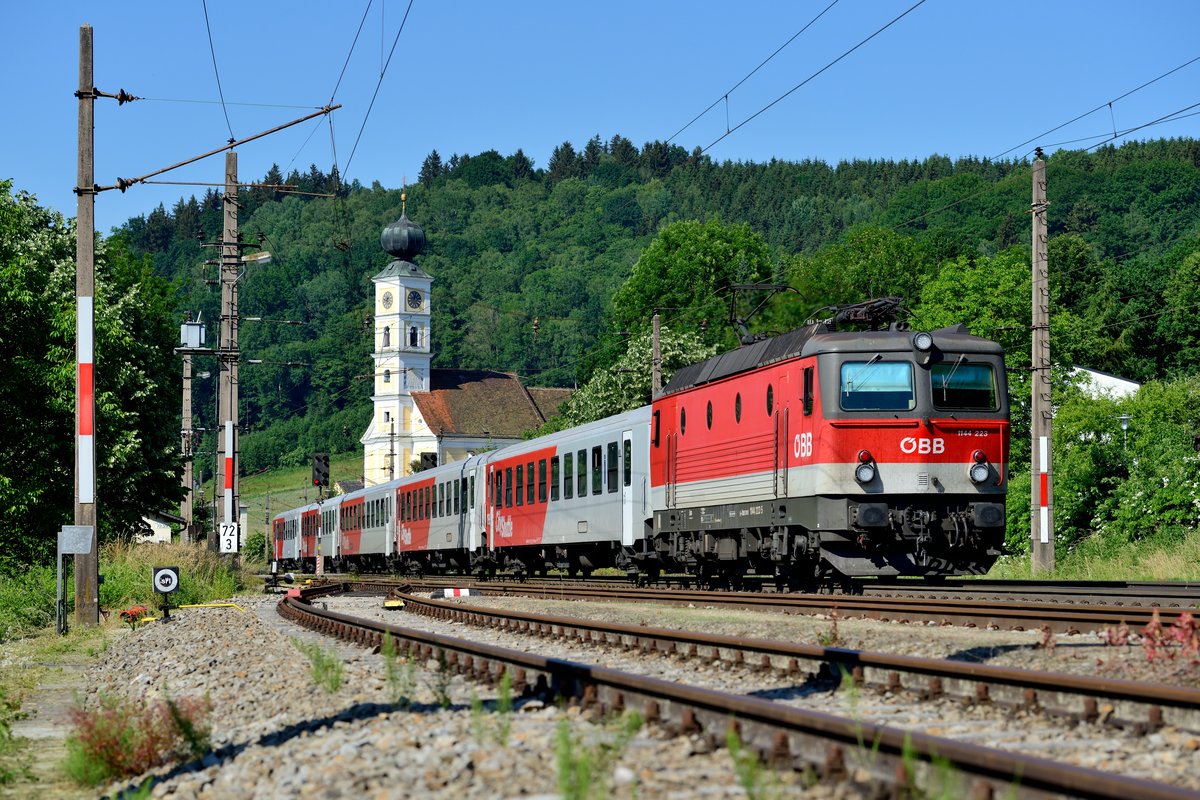 Das Motiv einmal aus der  Froschperspektive : Die gut gepflegte 1144.223 läuft mit ihrem REX 1785 nach Linz HBF in den Bahnhof Wernstein ein. Im Hintergrund die Pfarrkirche zum hl. Georg (12. Juni 2015).