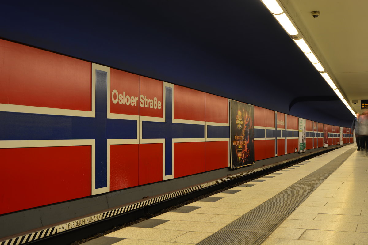 Ein Hauch von Skandinavien. Blick durch die U-Bahn Station Olsoer Straße.

Erste Planungen für einen Schnellbahnhof an dieser Stelle gehen auf das Jahr 1907 zurück. Die damaligen Planungen sahen an dieser Stelle jedoch einen Hochbahnhof vor. Die AEG-Schnellbahn A.G. plante eine Hoch- und Untergrundbahn von dort nach Rixdorf. Infolge der wirtschaftlich schwierigen Zeit nach dem Ersten Weltkrieg wurde die AEG-Schnellbahn A.G. jedoch 1923 liquidiert. Die Stadt Berlin, die den Bau 1926 fortsetzte, verkürzte die Streckenführung bis zum Bahnhof Gesundbrunnen, sodass an der Osloer Straße vorerst kein Schnellbahnhof entstand.

Im Jahr 1955 beschloss der Berliner Senat ein U-Bahn-Ausbauprogramm (den ersten „200-Kilometer-Plan“), der erstmals die neue U-Bahn-Linie G, die heutige U9, berücksichtigte. Dieser Plan sah auch bereits für einen Ausbau der Linien U8 und U9 nach Norden den Kreuzungspunkt unter der Osloer Straße vor.

Die ersten U-Bahn-Bauarbeiten begannen im Bereich der Osloer Straße im Jahr 1973. Als Kreuzungsbahnhof wurde ein unterirdischer Turmbahnhof errichtet. In halber Tieflage befindet sich ein großzügig angelegtes Zwischengeschoss mit zahlreichen Zugängen auf die verschiedenen Straßenabschnitte. Eine Ebene tiefer befindet sich der Bahnsteig für die U9, in zweieinhalbfacher Tieflage der 18,5 Meter breite Mittelbahnsteig der U8.

Wie für damals nahezu alle Stationen, war auch hier Rainer G. Rümmler für die Gestaltung zuständig. Beide Bahnsteige sehen sich zum Verwechseln ähnlich, die Hintergleiswände sind mit norwegischen Fahnen verziert. Die restlichen Wände wurden durch damals moderne, silberne Metallbleche verkleidet. Die Deckenfarbe war ursprünglich auf den beiden Bahnsteigen unterschiedlich, bei der U8 war sie gelb, bei der U9 blau. Heute sind beide Decken weiß gestrichen. Zum neuangelegten Bahnhof gehörte auch ein Verbindungstunnel zwischen den beiden U-Bahn-Linien. Die U8 wurde dadurch betrieblich unabhängiger, da Betriebsfahrten das Territorium der DDR, unter dem die U8 im Transit hindurchfuhr, umfahren konnte.

Der Bahnsteig der U9 wurde am 30. April 1976 eröffnet, der der U8 über ein Jahr später am 5. Oktober 1977. Am 27. April 1987 wurde die Verlängerung der U8 nach Norden bis Paracelsus-Bad eröffnet. Für die U9 ist der Bahnhof Osloer Straße der Endbahnhof geblieben. Verlängerungspläne wurden mittlerweile aufgegeben.

Einen Aufzug erhielt der U-Bahnhof Osloer Straße am 22. Juni 1990. In den Jahren 2003 und 2004 erfolgte eine Sanierung des U8-Bahnsteigs. Die silbernen Metallplatten wurden gegen dunkelblaue, der Linienfarbe entsprechende, eingetauscht. Der Boden wurde mit Granitfliesen gestaltet und ein Blindenleitsystem installiert. Eine nahezu gleiche Sanierung erfolgte darauf beim U9-Bahnsteig; hier ließ die BVG die silbernen Metallplatten gegen dunkelrote Emailleplatten austauschen. Auch auf diesem Bahnsteig wurde ein Blindenleitsystem eingerichtet. 

Berlin Gesundbrunnen 03.01.2018


Seite „U-Bahnhof Osloer Straße“. In: Wikipedia, Die freie Enzyklopädie. Bearbeitungsstand: 11. August 2017, 21:48 UTC. URL: https://de.wikipedia.org/w/index.php?title=U-Bahnhof_Osloer_Stra%C3%9Fe&oldid=168080682