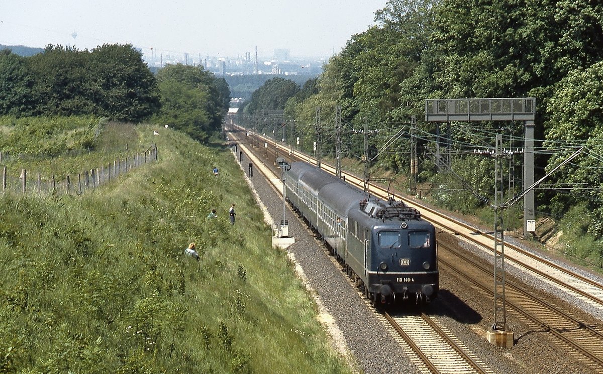 Im Juni 1987 fährt 110 148-4 die Steigung von Erkrath nach Hochdahl hinauf, der Blick von hier oben reicht an diesem Tag bis Düsseldorf. Die Rampe von Erkrath nach Hochdahl wurde 1841 in Betrieb genommen. Auf ca. 2,5 km überwindet die Bahn einen Höhenunterschied vom 82 m. Zur Zeit der Eröffnung mussten die Züge zunächst mittels einer stationären Dampfmaschine in Hochdahl an einem Seil gezogen werden. Später wurde die Dampfmaschine durch eine Umlenkrolle ersetzt und talwärts fahrende Lokomotiven oder Züge unterstützten die bergwärts fahrenden mittels Seil. Erst 1926 wurde dieses System aufgegeben, bis zur Elektrifizierung waren bis zu drei Dampflokomotiven notwendig, um schwere Züge vergwärts zu befördern. Die Herren, die den Hang bevölkern, warteten übrigens auf einen Dampfzug, dem ersten, der seit dem unsäglichen  Dampflokverbot  der DB hier unterwegs war.