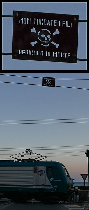 Nicht der Zug sondern das darüber hängende Schild im Fokus. Über dem Bahnübergang von Borgio Verezzi hängt ein Schild, das mit dem entsprechenden Gefahrensymbol darauf hinweist, die Leitung nicht zu berühren - Lebensgefahr. Aufnahme entstand am 30.10.2017.