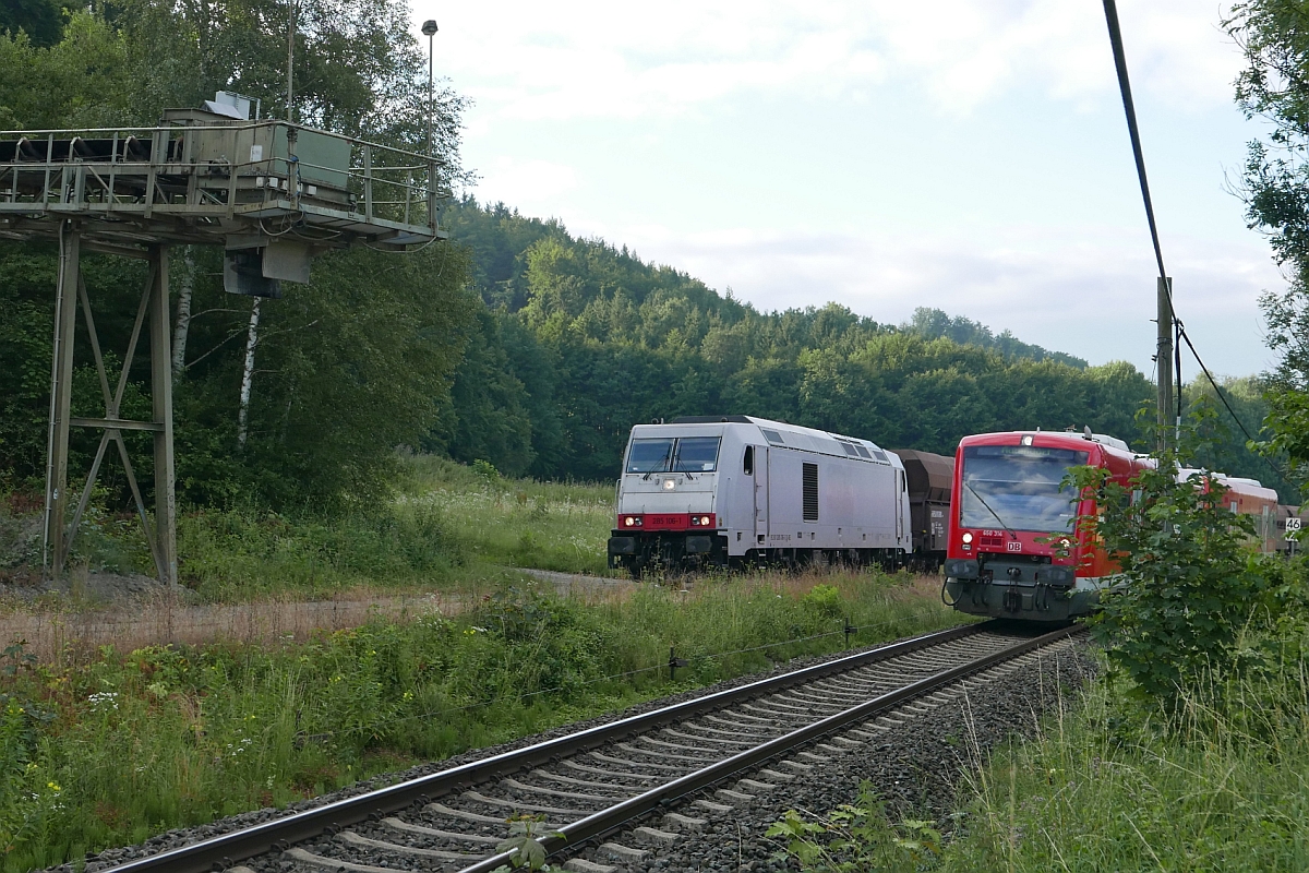 RB 22838 von Hergatz nach Aulendorf fährt am 'Roßberger Kieszug' vorbei, der mit 285 106-1 und Schüttgutwagen der Gattung Falns-x auf dem Ladegleis zur Beladung bereitsteht (Roßberg, 13.07.2017).