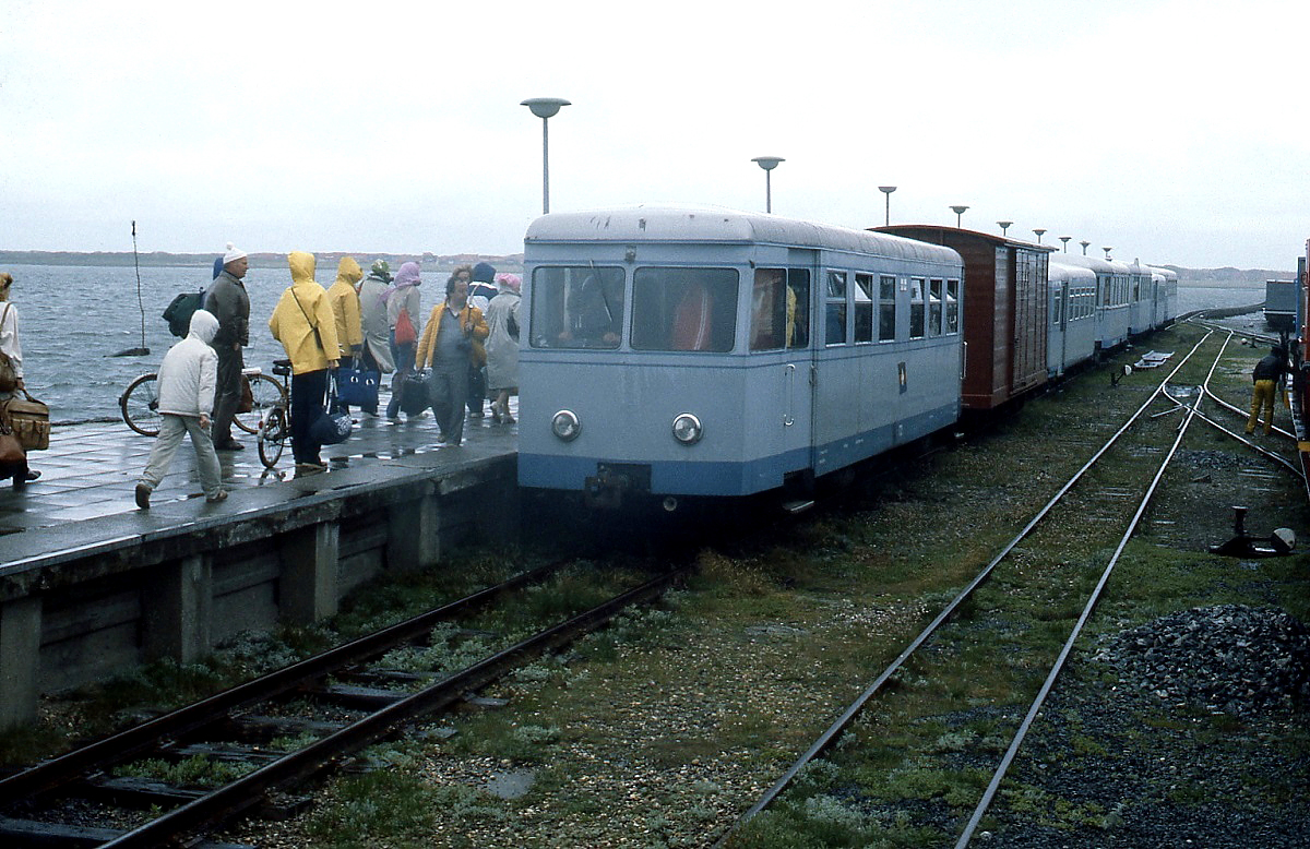 Von ziemlich unfreundlichem Wetter wurden die Urlauber an diesem Maitag 1981 auf der Insel Juist empfangen. Kein Wunder, dass sie es (bis auf den Fotografen) eilig hatten, in die Wagen der Inselbahn zu kommen, die sie über die Pfahlbaustrecke durch das Watt in den Ort bringen wird. T 2 am Zugschluss ist schon ziemlich gut besetzt. Nach dem Bau eines ortsnahen Hafens wurden Anleger und Strecke überflüssig und die Inselbahn 1982 stillgelegt. T 2 blieb aber erhalten und ist heute im Besitz des DEV (Bruchhausen-Vilsen).