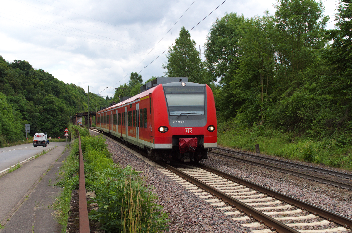 Zwischen Saarbrücken und Saarburg verläuft meist parallel zur Bahnstrecke 3230 die ehemalige Bundesstrasse 51. 425 129 ist als RB als Verstärker nach Saarhölzbach unterwegs. Gleich wird der Bahnhof Völklingen erreicht. Hier an der Luisenthaler Enge ist nur Platz für die Bahn und die Straße zwischen Felsen und der Saar. Rechts erhöht verläuft die Trasse der ehemaligen Köllertalbahn Völklingen - Lebach. Die Köllertalbahn überquerte auf der Brücke im Hintergrund die Saarstrecke und verschwand dann im Heidstock Tunnel. 04.07.2013