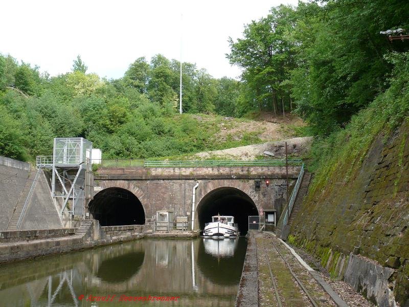 Blick auf die Tunnelportale der Eisenbahnstrecke und des Rhein-Marne-Kanals an der westlichen Seite der Vogesen. Zur berwindung des Gebirges fhren hier beide Verkehrswege durch je einen Tunnnel. Kanaltunnels gibt es zwar in Frankreich noch weitere, aber die Situation der parallelen Lage von Bahn, Kanal und ehemaliger Treidelbahn ist schon einzig. Hier mal der Beweis, da auch der Wasserweg befahren wird, wenn auch praktisch nur noch von Freizeitkapitnen und Hausbooten. Die kommerzielle Kanalschifffahrt rentiert sich durch  die vielen zeitraubenden und kleinen Schleusen und die geringe mgliche Tragkraft der Kanalschiffe seit lngerem nicht mehr.
 brigens ist fr die geplante TGV-Neubaustrecke ein Vogesenbasistunnel geplant, der auch in dieser Gegend beginnen soll.

13.05.2007 Arzviller-Tunnel