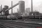 Mit einem leeren Kohlenzug fährt eine 215 vorbei an einer Dampflokomotive der Grube Anna in den Bahnhof Alsdorf ein (März 1978).