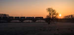 Sonnenaufgang an der Mainbrücke zwischen Kostheim und Bischofsheim. Während die Wiese noch von Reif überzogen ist und im Hintergrund die ersten Flugzeuge vom Frankfurter Flughafen starten, passieren zwei Traxx-Lokomotiven mit einem Güterzug das Bauwerk. (15.02.2017)