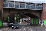 Hinter der Bahnbrücke kommt ein Tram ins Blickfeld -    Eine Urbos-Tram auf der Linie 4 am Konturplatz in Freiburg kurz vor der Unterführung unter der Rheintalbahn.