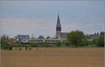 Bahn im Elsass. Bei aufziehendem Schlechtwetter fhrt ein TER an Raedersheim vorbei. Vor der markanten Kirche passt auch ein markanter Steuerwagen ins Bild. April 2017.