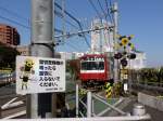 Die Stammstrecke des Keihin Kyûkô (Keikyû)-Konzerns: Die Strassenkreuzung vor Tokyo-Shinagawa. Auf dem Schild steht:  Sobald die Warnglocken ertönen, bitte nicht in den Bahnschrankenbereich hineintreten.  Im Hintergrund fährt Zug 827 Richtung Miura-Halbinsel aus. 28.Oktober 2011. 
