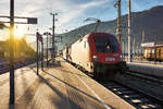 Während die Morgensonne in den Bahnhof Spittal-Millstättersee hereinstrahlt, durchfahren 1016 002-8 und 1144 228-4 mit einem Güterzug in Richtung Tauern den Bahnhof.