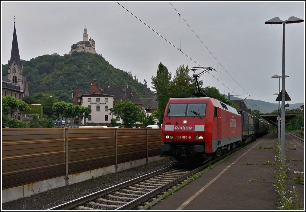 - UNESCO Welterbe Oberes Mittelrheintal - Im strmenden Regen durchfhrt am 25.06.2011 ein Gterzug den Bahnhof von Braubach, whrend hoch oben die Marksburg zu sehen ist. Sie steht auf einem Schieferkegel in 160 Meter Hhe und ist die einzige nie zerstrte mittelalterliche Hhenburg am Mittelrhein. Der Bau entstand zum Schutz und zur Verwaltung Braubachs und diente anfnglich auch als Zollburg. Mit ihrem heutigen Aussehen gilt sie als die mittelalterliche Burg schlechthin und ist Vorbild fr viele Phantasie-Burgen, insbesondere als Spielzeug-Burganlagen. (Hans) 