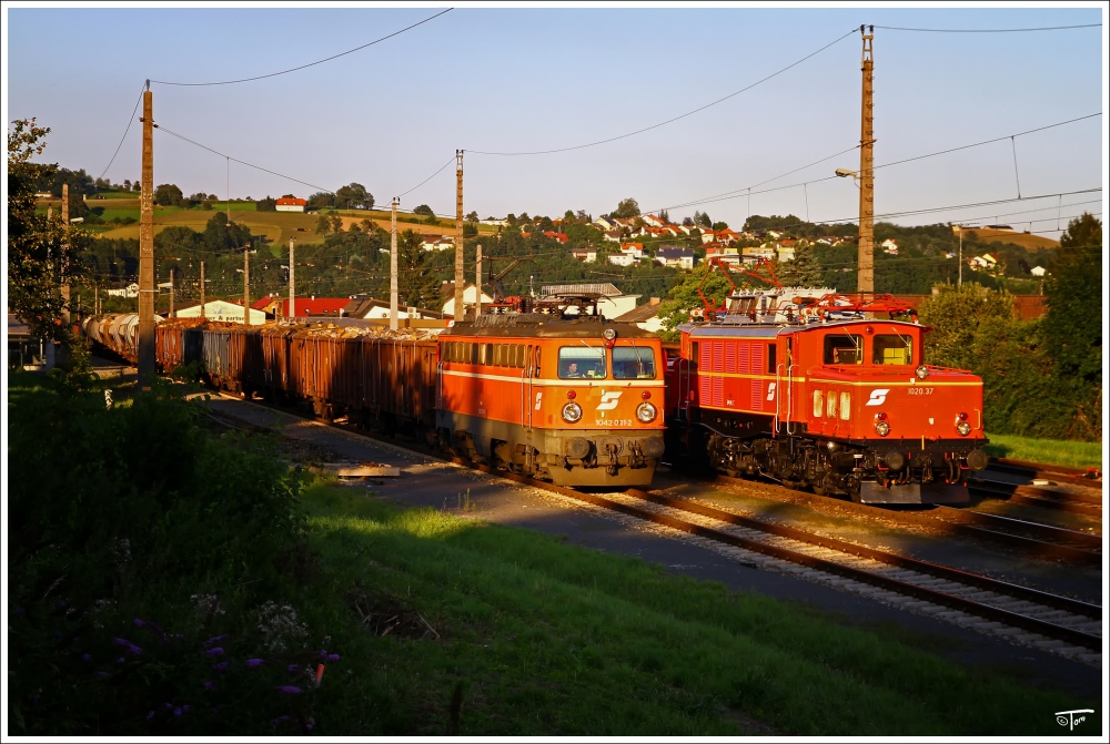 1020 037 steht mit 94699 (Plan 47541)von Summerau nach Linz-Stahlwerke im Bahnhof St Georgen an der Gusen, während im Vordergrund 1042 041 mit einem Holzzug vorbeifährt. 
9.8.2010