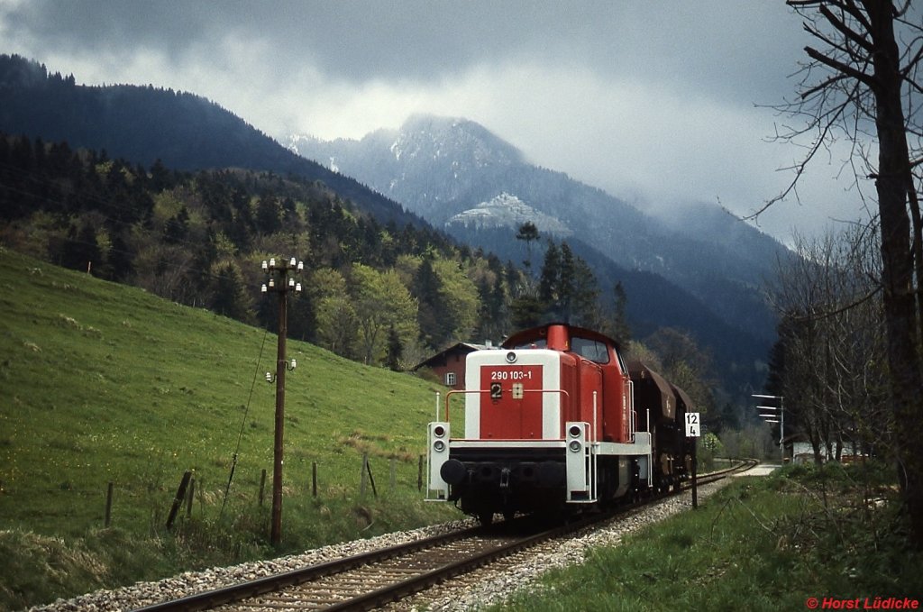 290 103-1 durchfhrt im Frhjahr 1989 mit zwei Schotterwagen den Haltepunkt Geitau in Richtung Schliersee. Der Bahnsteig lag damals noch vor dem im Hintergrund rechts hinter den Bumen erkennbaren frheren Bahnhofsgebude, das allerdings schon lange in Privatbesitz war. Der neue Hochbahnsteig der Oberlandbahn befindet sich etwa in Hhe der Lok.