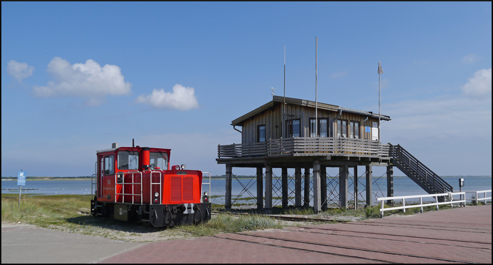 399 107-2 der Wangerooger Inselbahn setzt im Westanleger auf die andere Seite des Zuges um, nachdem sie viele Urlauber vom Inseldorf an den Hafen gebracht hatte. Das auf hohen Bohlen gebaute Haus gehört zum Jachthafen und stellt eine Grenzaufsichtsstelle dar, wenn man aus dem Ausland mit dem Schiff hergereist ist. 