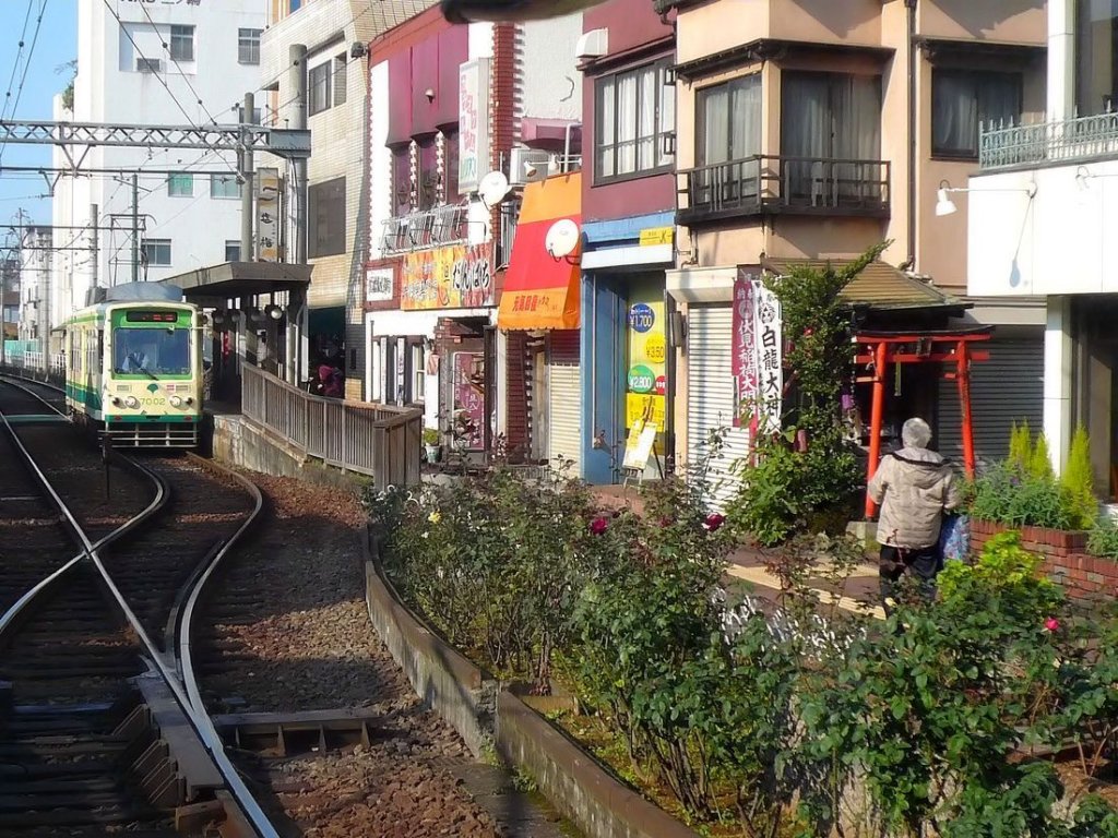 7002 heute: Am 28.November 2009 steht 7002 an der Entstation Minowabashi. Die aussteigenden Fahrgäste kommen an winzigen Geschäften, u.a. einem kleinen Restaurant und einem Friseurladen, sowie (rechts, durch rote Pfosten gekennzeichnet) einem ganz kleinen Götterschrein für die Fruchtbarkeitsgottheit Inari vorbei. Wagen 7002 hat einen Wagenkasten von ca.1977 und stammt im übrigen aus Bauteilen der alten Wagen Serie 7000 von 1954-1956. 