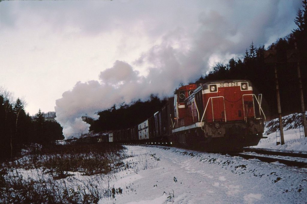 An einem eisigen Morgen schiebt die DE10 526 einen schweren Güterzug über den Jômon-Pass bei Engaru in Ost-Hokkaido. Das zarte Rosa der Morgensonne schimmert in den Dampfwolken der Zuglokomotive (D51 511). 29.Dezember 1971. SEKIHOKU-HAUPTLINIE
