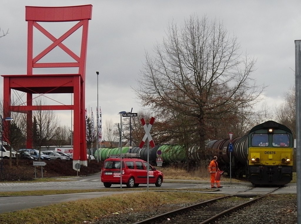 DE676 der ASCENDOS fhrt mit den leeren Kesselwagen am 08.03.2012 ber einen unbeschrankten und nicht mit Lichtzeichen ausgestatteten Bahnbergang, der von einem Bahnmitarbeiter gesichert wird. DE676 befindet sich auf der Rckfahrt von einem Heizlhndler im Industriegebiet Friedrichshafen nach Friedrichshafen Gbf.