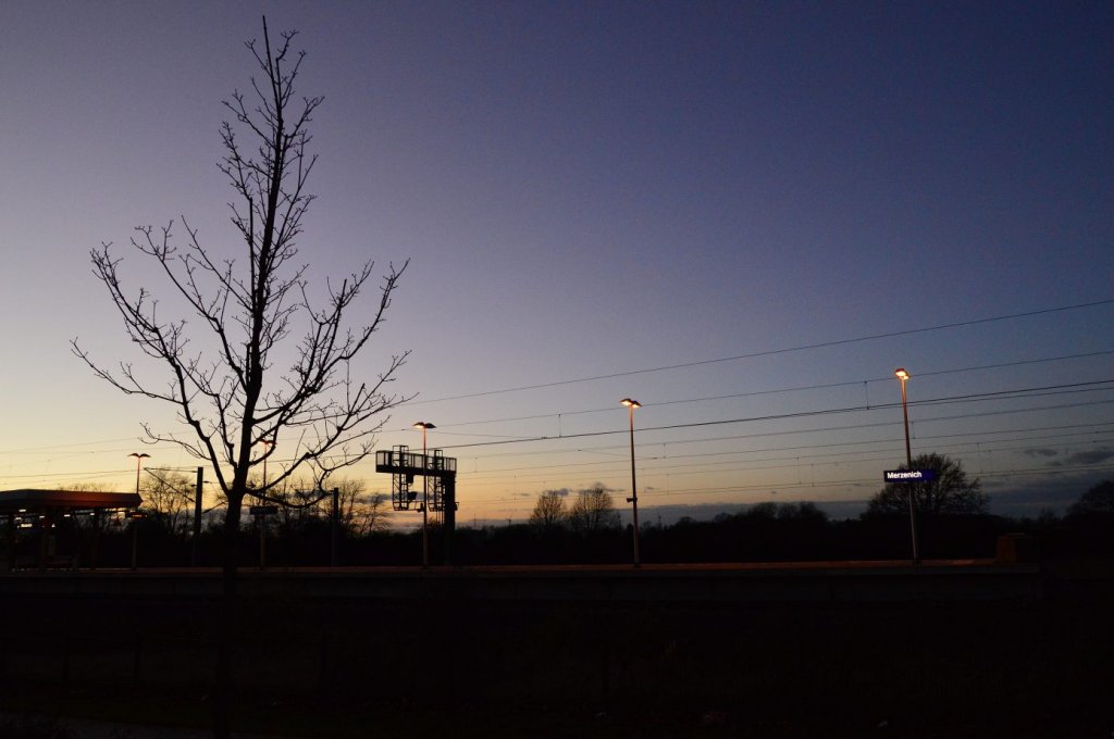 Dieses Bild zeigt den S-Bahnsteig in Merzenich beim Sonneuntergang am Sonntagabend den 25.11.2012 vor wenigen Minuten ist die Bahnsteigbeleuchtung eingeschlatet worden und die Ortsnamestafel leuchte nun jedem die Station.....im Hintergrund erkennt man die Signalbrcke mit den Ks-Signalen fr die in Richtung Aachen fahrenden Zgen des Schnellfahrabschnitts.