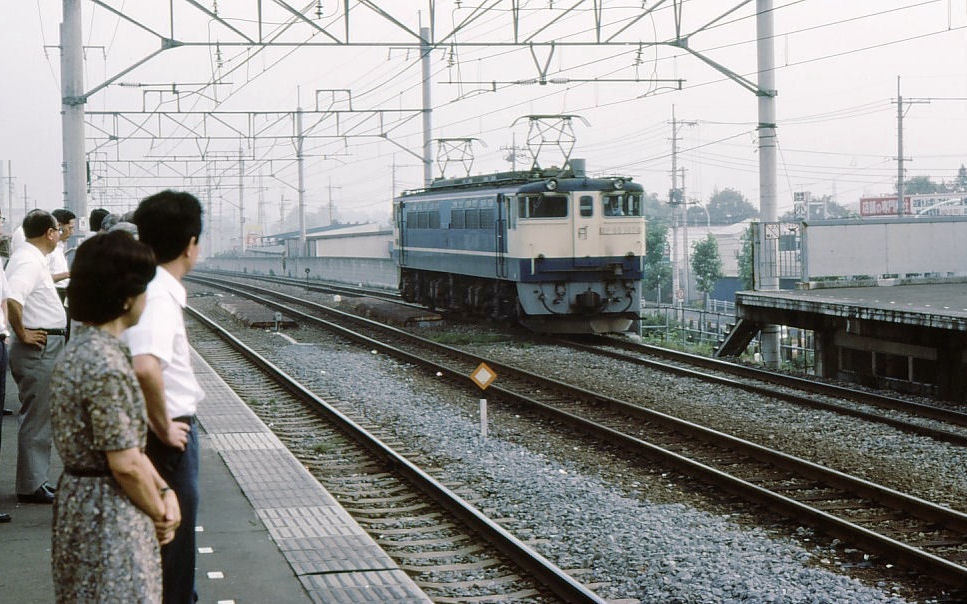 EF65: An einem unglaublich schwül-heissen Tag warten die Leute auf einen stickigen S-Bahnzug, während auf dem anderen Gleis die EF65 1074 vorbeidonnert. Tokyo-Nishi Kokubunji, 2.Juli 1979. 