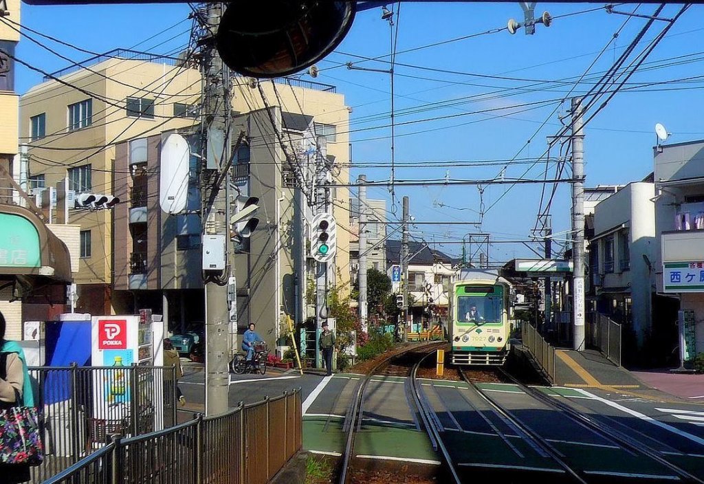 Einst und jetzt: über 30 Jahre sind seit der vorangehenden Aufnahme vergangen. Wagen 7027 ist den Berg heraufgekommen und hält an der Station Nishigahara Yonchôme. Das Masten- und Drahtgewirr ist noch da, nur moderner, der Gebäudeblock im Hintergrund hat die Farbe gewechselt, die kleineren Häuser sind neu. Auch Wagen 7027 trägt ein neues Farbkleid. 28.November 2009.