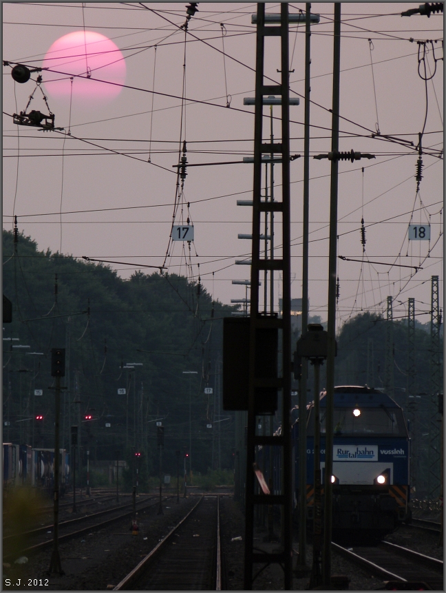 Glutrot und richtig fett stellt sich die Sonne als Motiv zur Schau. Die 
Vossloh Lok der Rurtalbahn wartet schon sehr lange auf ihr Ausfahrtssignal.
Happy Hour in Aachen West am 26.Juli 2012 Sptabends.