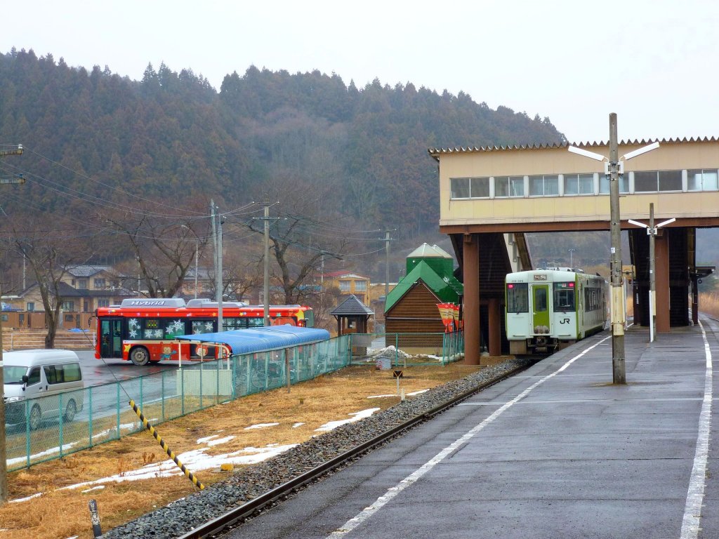 Hier in Yanaizu endet heute die Kesennuma-Linie; links ist der Bahnersatz-Bus (von JR Ost-Japan betrieben) zu sehen, der die Fahrgäste weiter in die Katastrophengebiete bringt. Der Bus ist in fröhlichem Leuchtendrot gestrichen und mit Bildern der verschiedenen Maskottchen und Figürchen versehen, welche die Stationen der Kesennuma-Linie markiert haben. Im Bahnhof steht der einzelne Triebwagen (KIHA 110-125), der das Reststück der Bahnlinie bedient; 15.Februar 2013. 