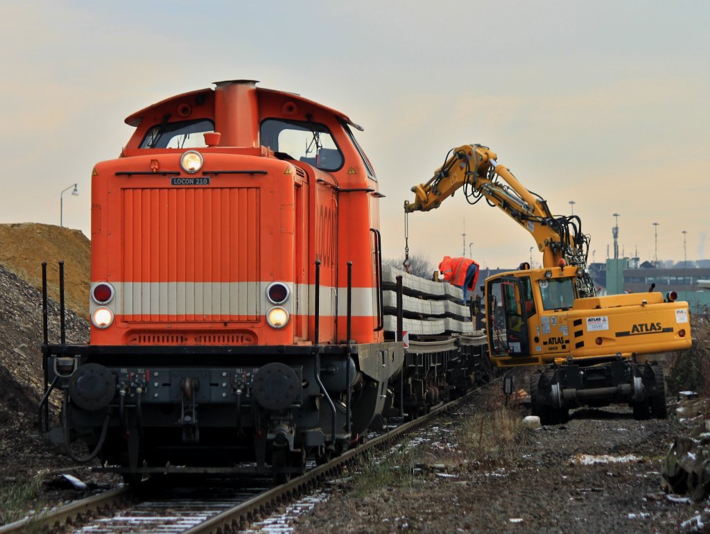 Locon 210 (212 275-2) steht am 13.12.2012 mit einem Schwellenzug in Aachen West und wird von einem Zweiwegebagger der Firma Balfour Beatty entladen.