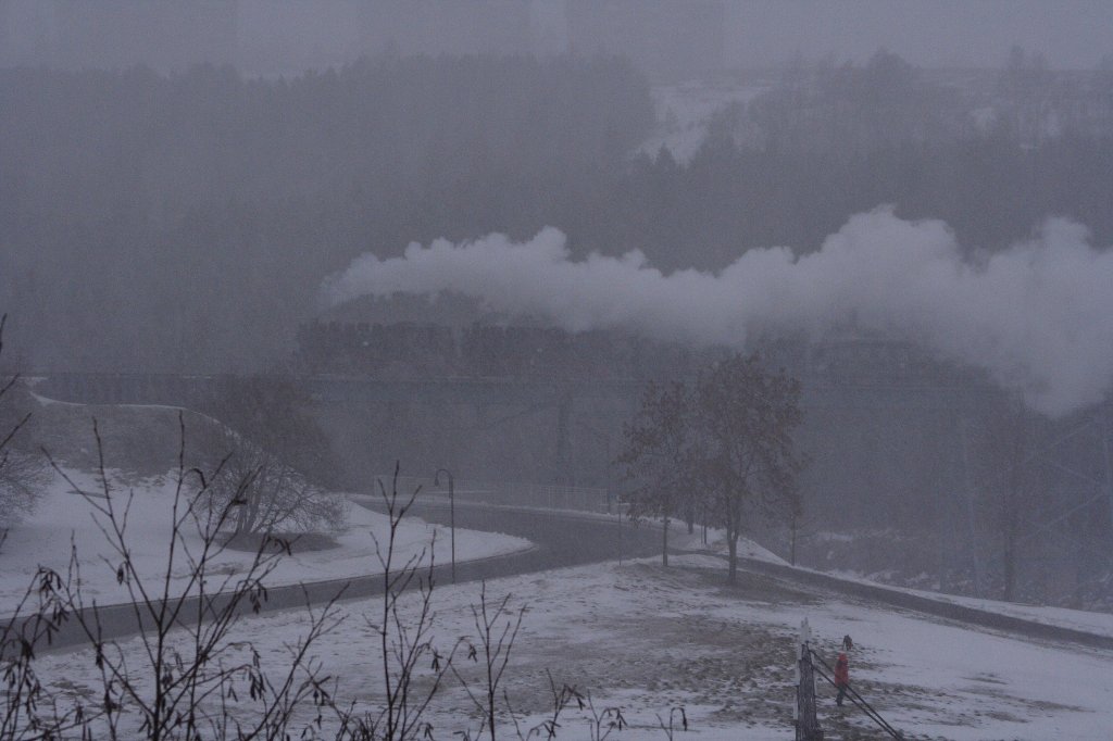 Mit viel Getse kmpfen sich am frhen Nachmittag des 30.12.2012 99 785 und 99 786 mit ihrem langen Personenzug P1005 aus Cranzahl, whrend eines heftigen Schneeschauers ber das Oberwiesenthaler Viadukt die Steigung zum Bahnhof hinauf. Die Entstehungsgeschichte fr dieses Bild war doch etwas kurios! Nachdem ich eine Stunde (!) nach dem geeigneten Aufnahmestandort gesucht hatte (ist gar nicht so leicht zu finden!) und endlich fndig geworden war, nutzte ich die verbleibende Zeit bis zur Ankunft des Zuges(ca. 30 min) fr ein paar Probeaufnahmen. Whrenddessen zog ber dem Fichtelberg eine schwarze Wolkenwand auf, welche nichts Gutes verhie! Folgerichtig setzte auch kurz vor Erscheinen des Zuges (er war bereits zu hren!) ein gewaltiger Schneeschauer ein. Wild entschlossen, dennoch ein Bild zu machen, harrte ich aber auf meinem Standort aus, wobei sich mein Erscheinungsbild in krzester Zeit grundlegend vernderte. Htte man mir einen Eimer auf den Kopf gestlpt, wre der Schneemann perfekt gewesen, denn die rote Nase hatte ich, wetterbedingt, ohnehin schon. Nachdem ich meine Aufnahmen gemacht und der Zug das Viadukt passiert  hatte, lie der Schneeschauer genauso pltzlich, wie er gekommen war, wieder nach, die Wolkendecke riss auf und blauer Himmel und Sonne kamen wieder zum Vorschein! Die Gedanken, welche mir dabei durch den Kopf gingen, sind fr eine Verffentlichung absolut ungeeignet, kann sich aber wohl jeder selbst vorstellen! ;-) Und das besonders, wenn man bedenkt, dass es an diesem Tag die einzige Chance war, die ich hatte, von diesem Zug eine Aufnahme zu machen! Ich hoffe, es ist aber trotz allem ein halbwegs interessantes Bild geworden. 