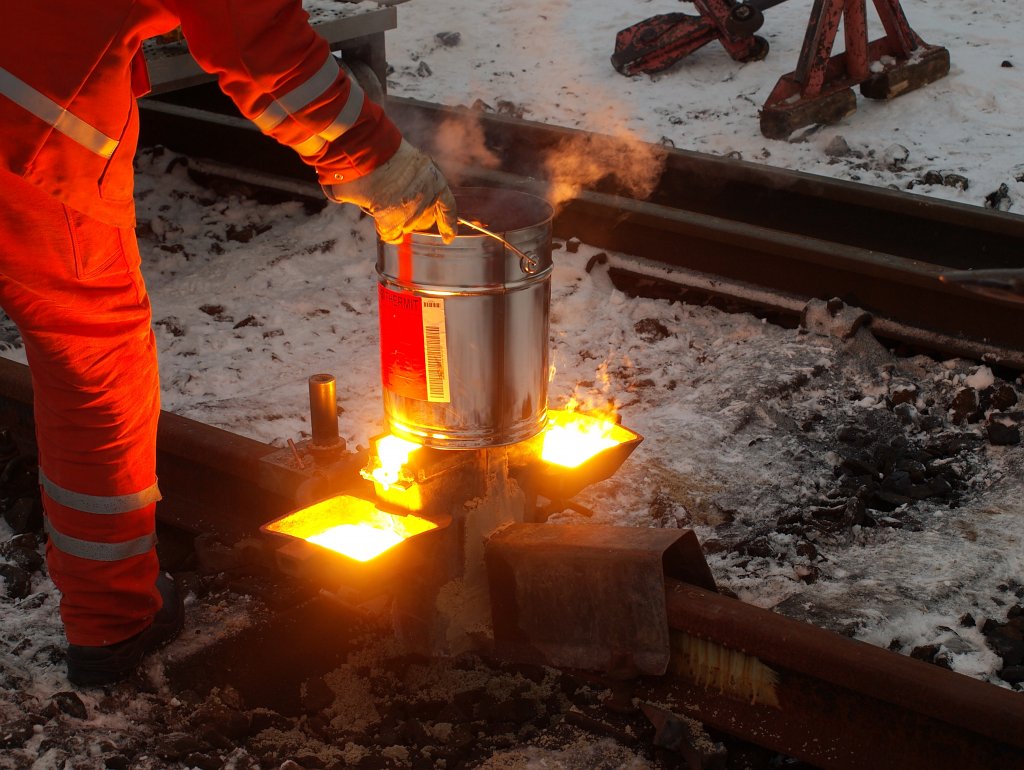 Schweieinsatz in Aachen West am 14.12.2010. Hier sieht man die Gussform und die Formhaltebleche, die mit einer Spannvorrichtung  an den Schienenkopf geklemmt sind. Die Form wird mit Sand gegen die Schiene abgedichtet. Die Schiene wird vor der Gussform zustzlich mit einem Blech geschtzt um eventuell daneben laufendes Metall fernzuhalten. Der Thermit-Tiegel wird  nun von der Gussform genommen, nachdem durch die chemische Reaktion eine Temperatur von ca. 2400C ereicht ereicht wurde und das flssiges Eisen in die Form und die leichtere Aluminium-Schlacke ber zwei berlufe in die Auffangschalen gelaufen ist. Anschlieend werden die Schlackeschalen, die Haltebleche fr die Form und die Schutzbleche fr die Schiene entfernt. Nach kurzer Zeit, wenn der Stahl soweit abgekhlt ist das er fest wird, kann die Form abgeschlagen und mit dem Abschergert der berstehende Thermit-Stahl am Schienenkopf beseitigt werden. Der Rest wird abgeschliffen und schon ist die Schiene wieder befahrbar.