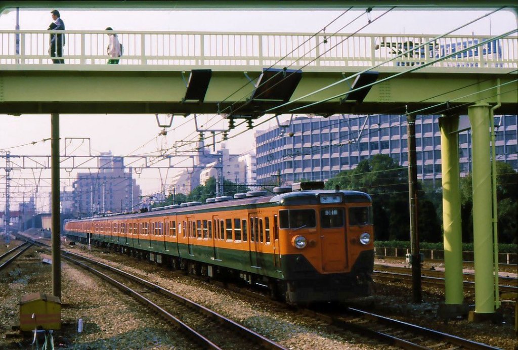 Serie 111 u.113: Die grn/orangen Zge (sie symbolisieren die Mandarinenplantagen zwischen Tokyo und Nagoya) waren fr den Lokalverkehr auf der Tkaid-Strecke entlang des Pazifiks zwischen Tokyo und Osaka bestimmt. Hier ist ein Zug mit Steuerwagen KUHA 111-85 in Shin-Osaka; ein Blick aufs Dach zeigt, dass noch keine Klimaanlage eingebaut ist. 27.Mrz 1986. 