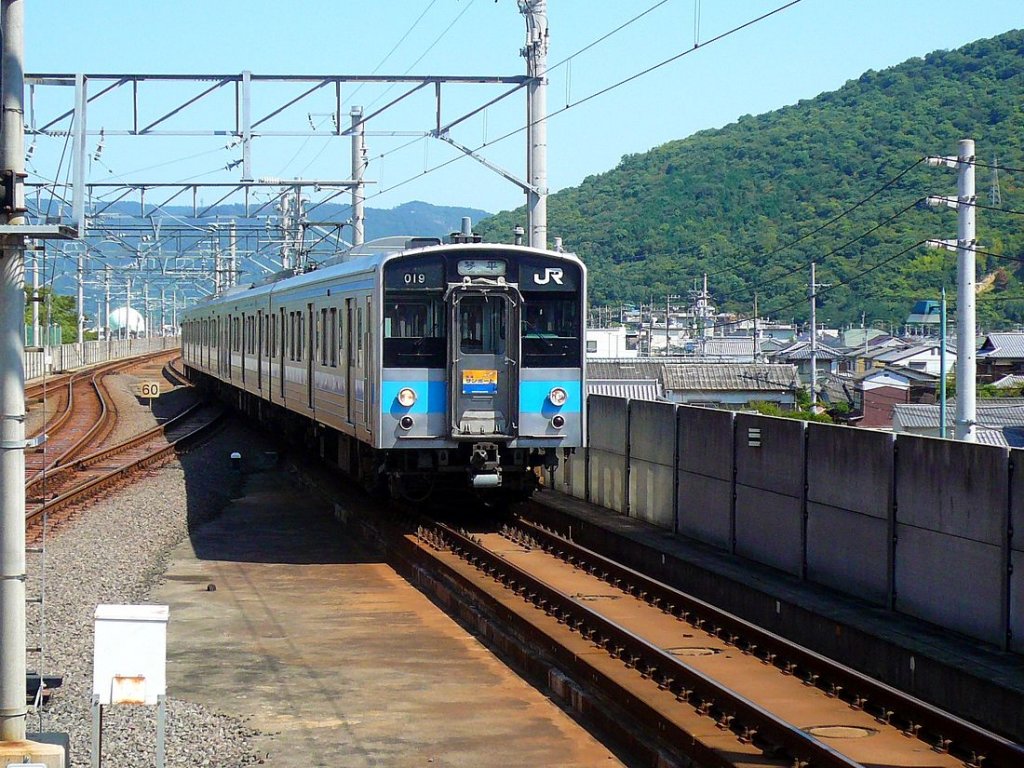 Serie 121: Ein Zug aus zwei Zweiwagen-Einheiten erreicht die Station Sakaide. Diese Station am shikoku-seitigen Ende der riesigen Inlandseebrücke (Seto Ôhashi), die die Landverbindung mit der japanischen Hauptinsel herstellt, wandelte sich nach deren Eröffnung 1988 in kürzester Zeit von einem schläfrigen Dorf in ein Industriezentrum. Der eintreffende Zug wird von Steuerwagen KUHA 120-19 geführt. 17.September 2009. 