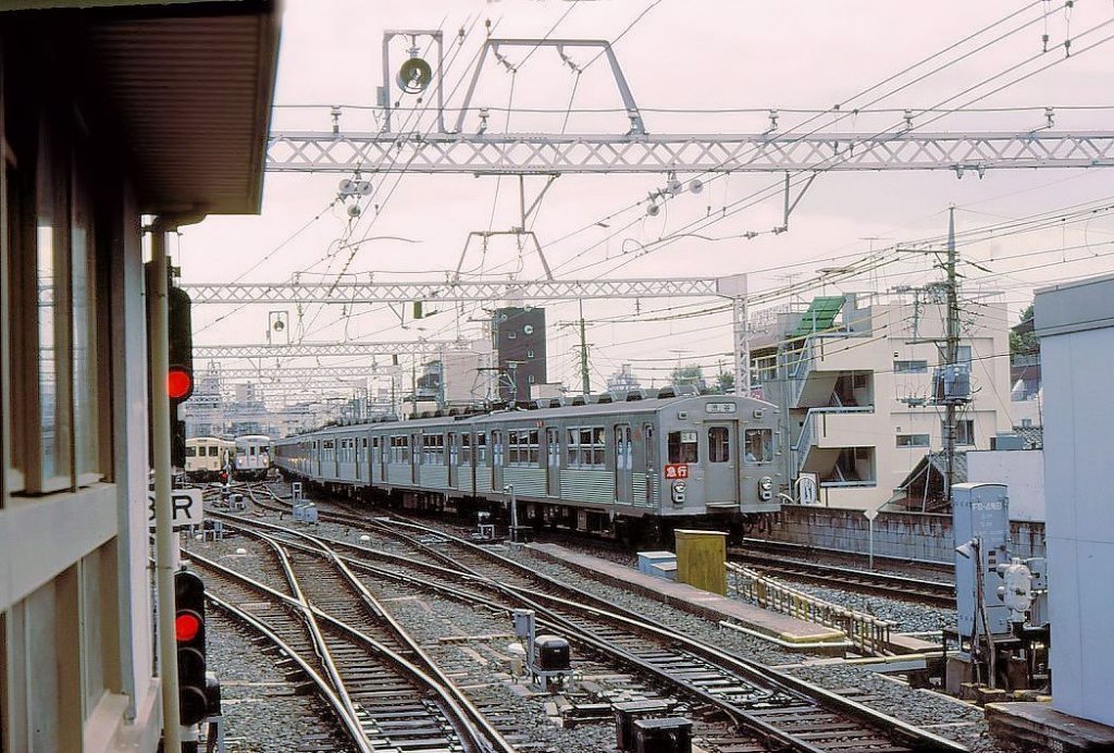 Tôkyû-Konzern, Serie 7000 / Tokyo Metro Hibiya-Linie: Diese Pionier-Züge mit Kasten aus rostfreiem Stahl (in Zusammenarbeit mit der amerikanischen Budd Co. gebaut) entstanden 1961-1966. Es waren die ersten Züge, die von einer S-Bahnstrecke her auf die Tokyo U-Bahn einfahren konnten. Die Serie bestand aus 134 Wagen; nach ihrem Ausscheiden 1991 gelangten sie in ganz Japan an verschiedene Privatbahnen, etwa zur Mizuma-Bahn oder zu den Fukushima Verkehrsbetrieben. Im Bild: Einfahrt des Zuges 7001 in Tokyo-Naka Meguro; im Hintergrund zwei Züge, die auf Einfahrt auf die Hibiya-Linie der Tokyo U-Bahn warten (weiss ein Zug des Tôbu-Konzerns, silbrig ein U-Bahnzug). 2.Juli 1979. 