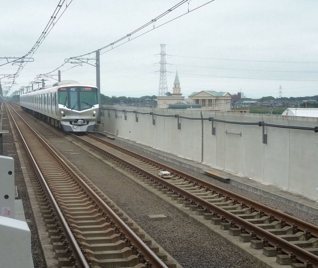 Tsukuba Express Serie 1101: 14 6-Wagenzge (1101-1114), ab 2003 fr den Gleichstromabschnitt von Tokyo bis Moriya (km 37,7) gebaut. Bild: Zug TX 1113 in Kashiwa-no-ha Campus, 25.Juni 2011. Im Hintergrund die rtliche Hochzeitszeremonienhalle. 
