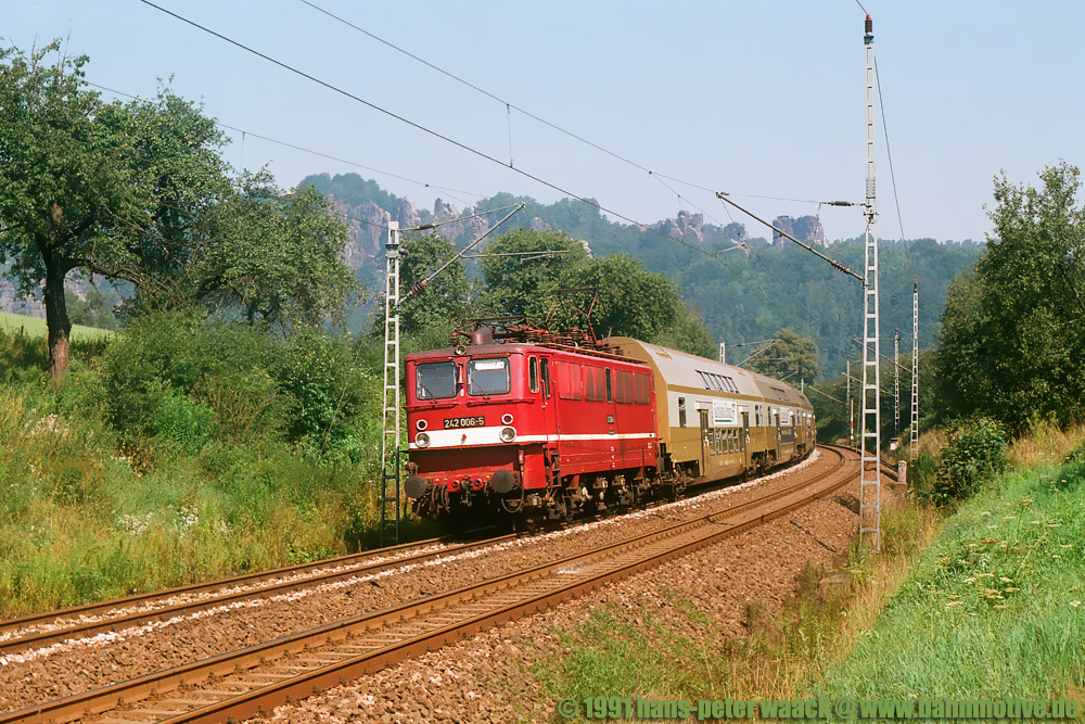 Zwischen Kurort Rathen und Knigstein im Elbtal kommt 242 006 am 07.08.91 mit einer S-Bahn S1 gefahren. Im Hintergrund die Felsen des Rathener Gebietes.