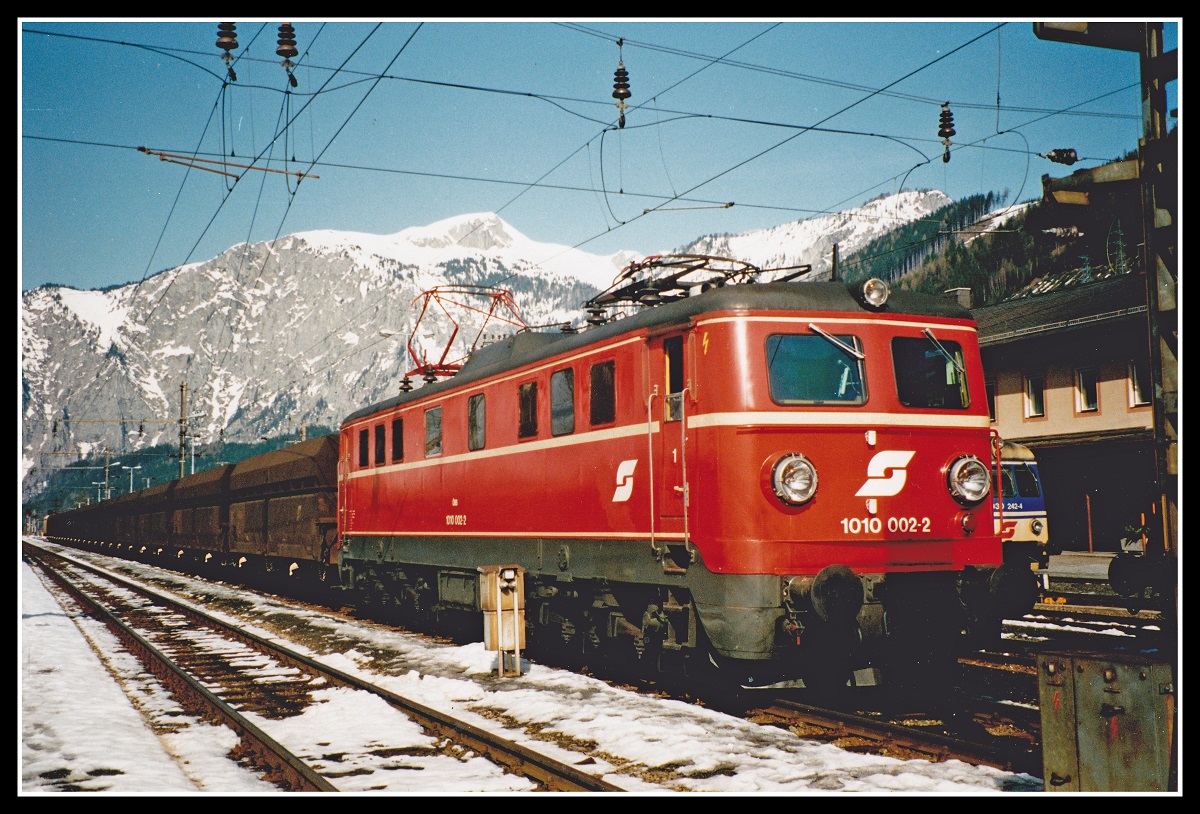 1010.002 ist am 27.02.1992 soeben mit einem Erzleerwagenzug in Eisenerz angekommen. Rechts neben der Lok ist der Triebwagen 4030.242 zu erkennen der in wenigen Minuten als Regionalzug nach Hieflau abfährt. Im Hintergrund ist die Seemauer und der Hochblaser ,1771 Meter hoch zu erkennen. 
