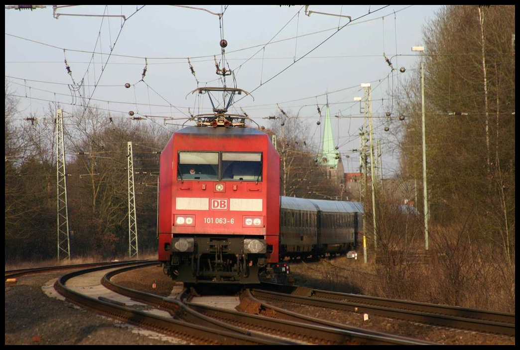 101063-6 hat gerade den Bahnhof Hasbergen durchfahren und wechselt hier am westlichen Bahnhofskopf die Gleisseite. Der von ihr gezogene Intercity ist am 20.3.2005 unterwegs nach Münster in Westfalen. Im Hintergrund ragt die Kirchturmspitze der evangelischen Kirche über die Bäume.