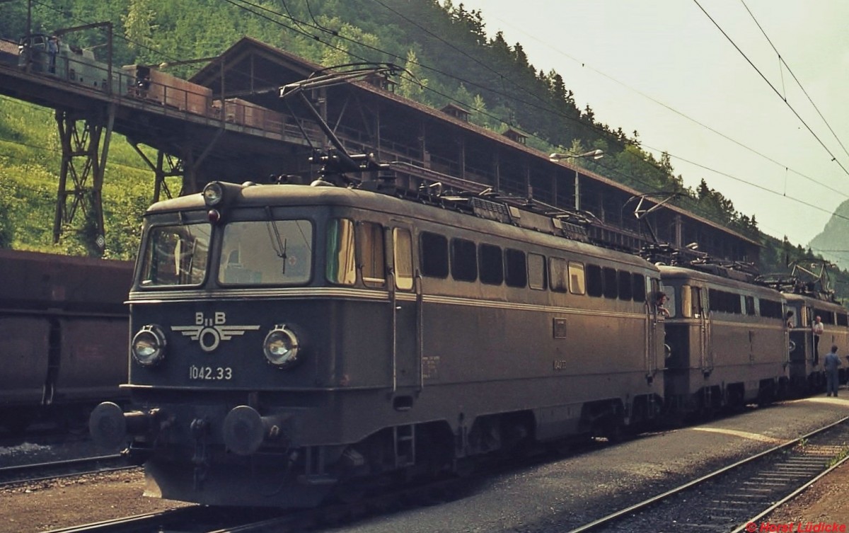 1042.33 und zwei weitere 1042 im Juni 1974 im Bahnhof Eisenerz. Die Lok besitzt noch Alu-Zierstreifen. Im Hintergrund die lngst verschwundene Erzverladeanlage mit einem Zug der schmalspurigen Huntslaufbahn.