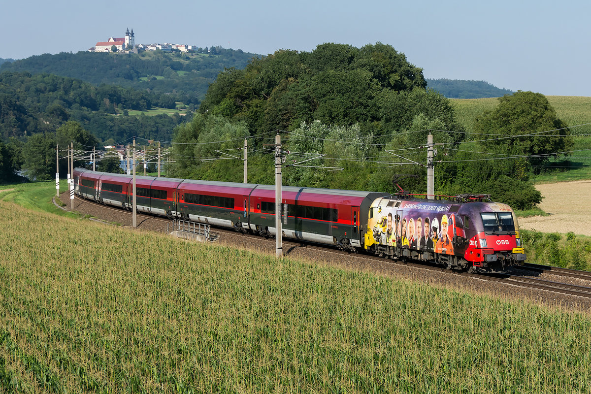 1116 153  ÖAMTC  mit railjet 742, unterwegs auf der alten Westbahnstrecke am südlichen Donauufer zwischen Pöchlarn und Ybbs/Donau bei Krummnussbaum, ein nicht alltägliches Bild. Diese Umleitung erfolgt aufgrund von Bauarbeiten, das Foto entstand am 08.08.2020.

Auf der gegenüber liegenden Seite der Donau befindet sich die Wallfahrtskirche Maria Taferl.