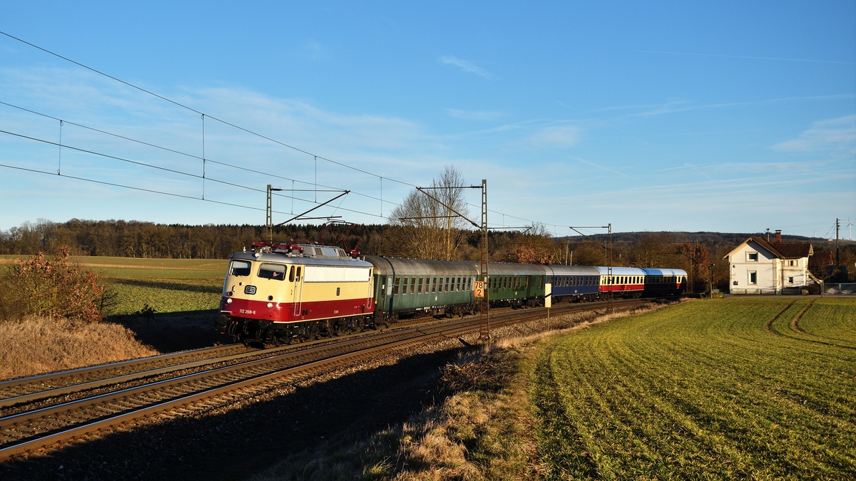 112 268 von TRI bespannt am 22. Februar 2020 den Sonderzug DPE 24659 der Ulmer Eisenbahnfreunde von Stuttgart nach Berchtesgaden. Erst seit wenigen Minuten liegt die Strecke kurz hinter Westerstetten im Licht, als der Zug die Stelle passiert.
