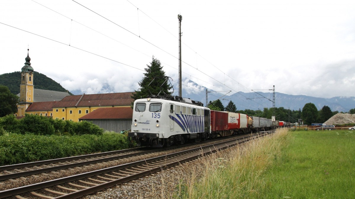 139 135-8 von Lokomotion durchfährt am 24.6.2014 mit einem KLV das Inntal bei Niederaudorf. Hier kurz nach dem BÜ mit dem Kloster Raisach. Im Hintergrund liegt das Kaisergebirge in dicken Wolken.